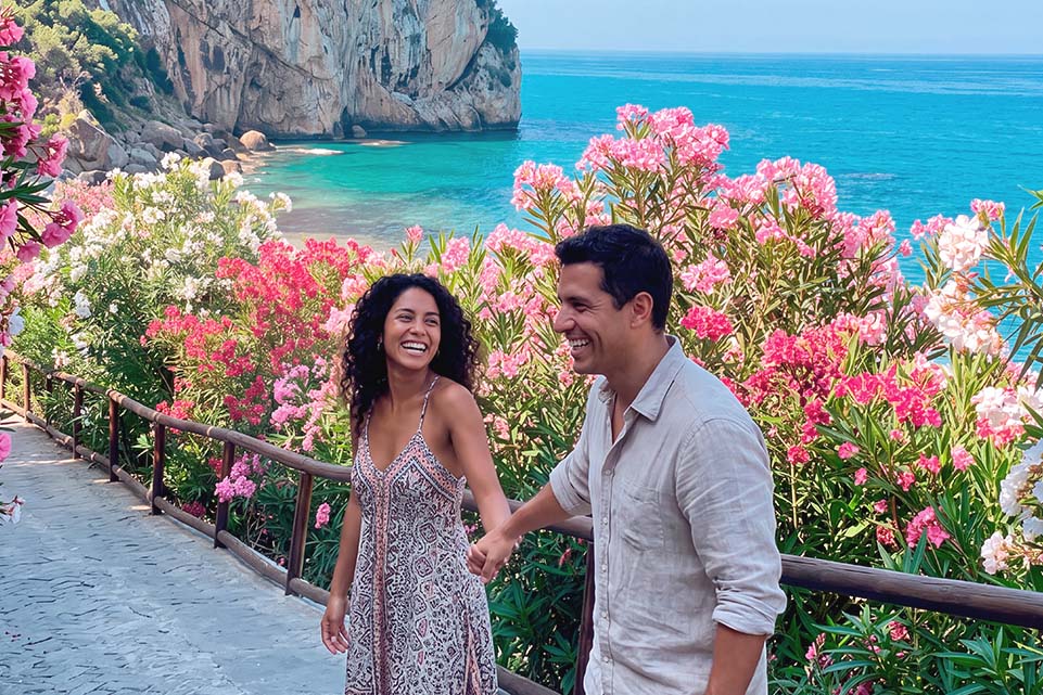 Pareja sonriente de la mano entre flores rosas, frente a una cala de agua turquesa. Imagen para Dinero de X Vida octubre 2026.